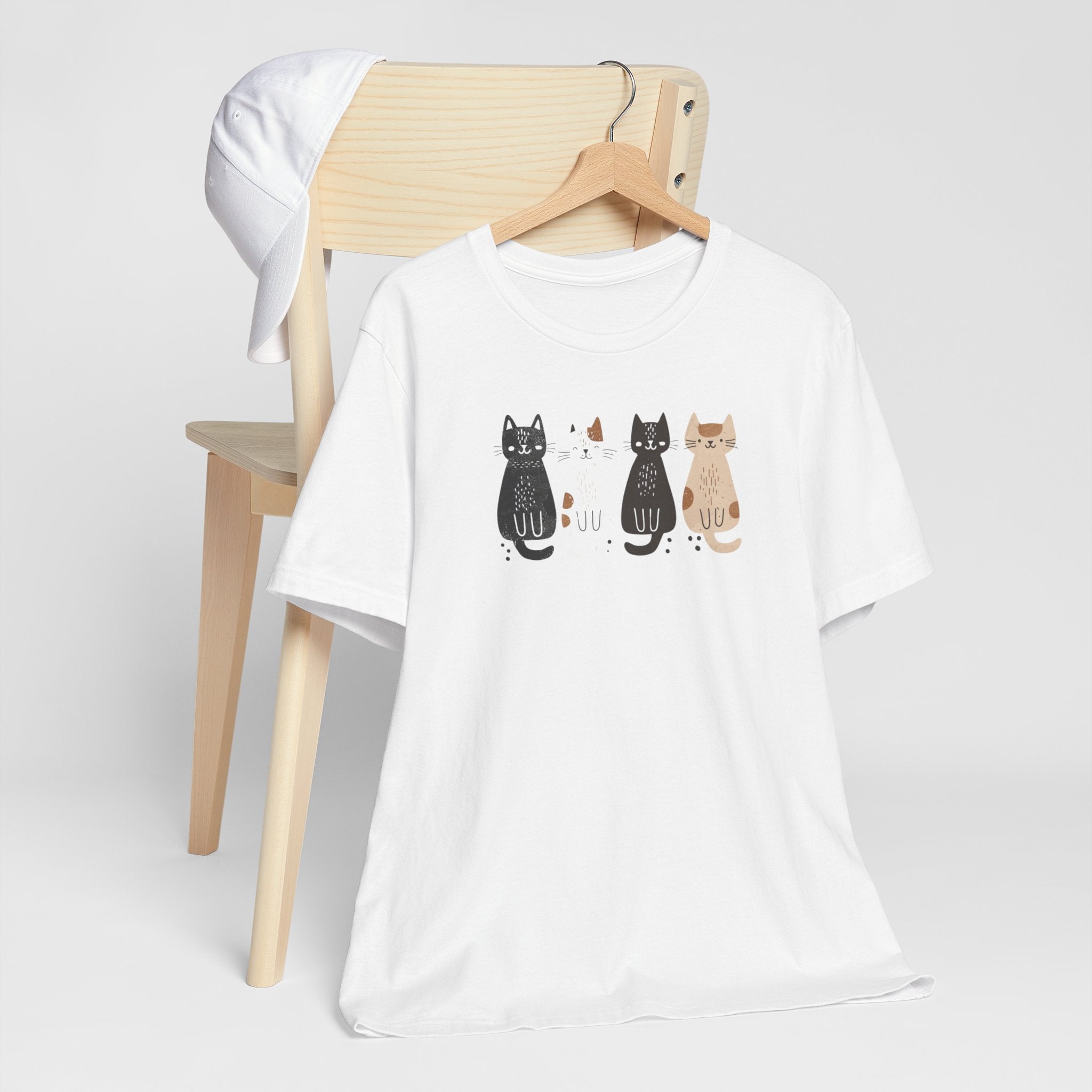 A white t-shirt featuring a graphic design of four cute cats in a lineup, each in different colors such as black, white, brown, and tan.