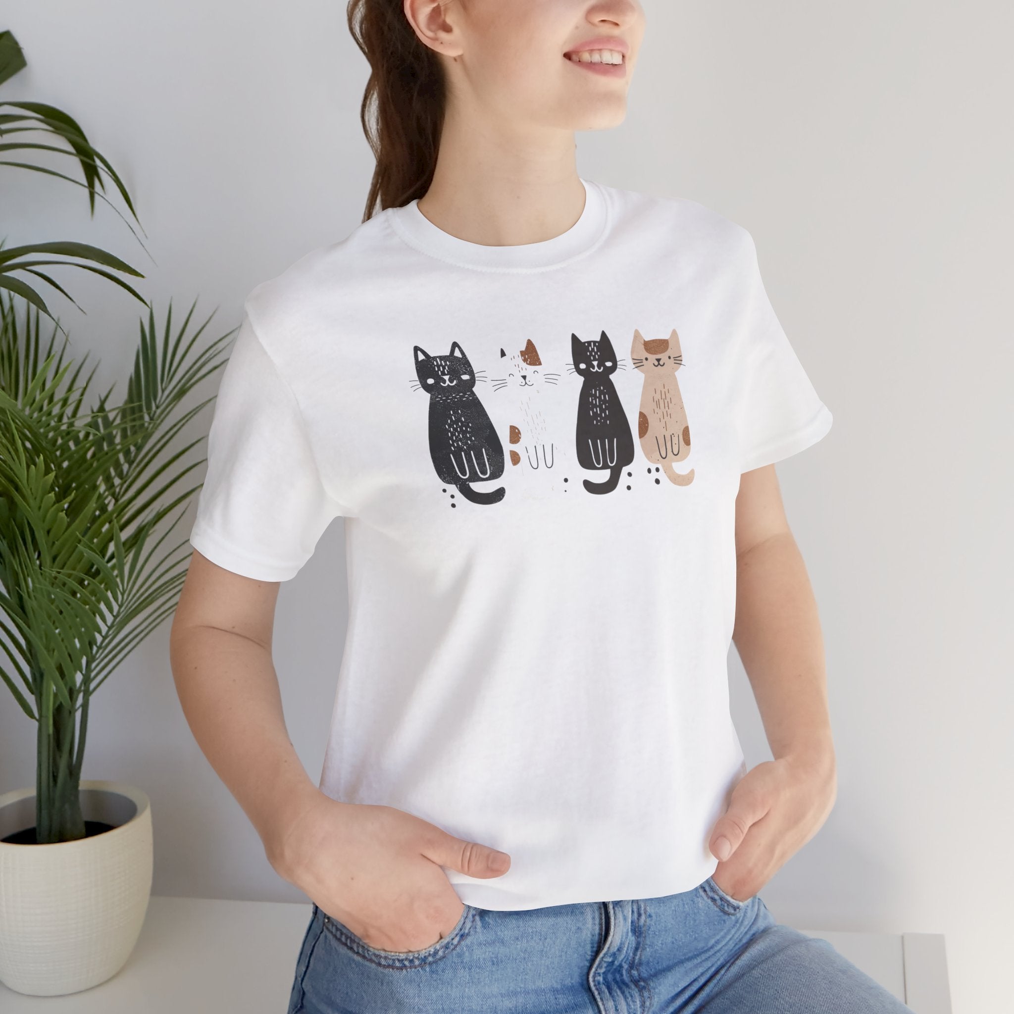 A white t-shirt featuring a graphic design of four cute cats in a lineup, each in different colors such as black, white, brown, and tan.