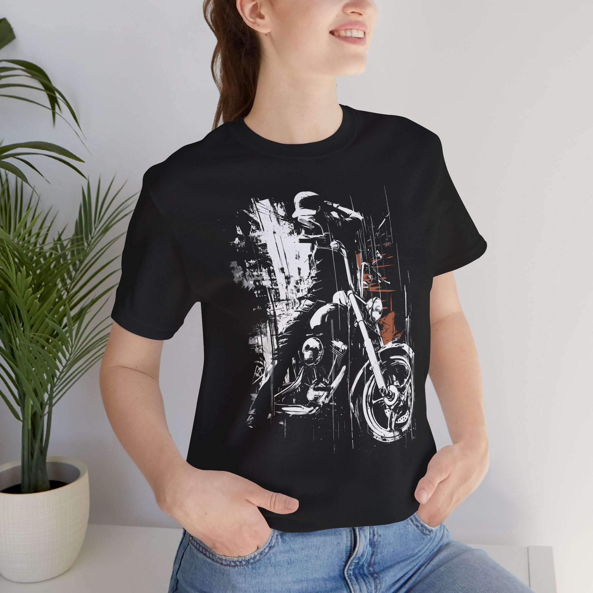 Person wearing a black t-shirt with a motorcycle graphic design, standing indoors next to a plant.
