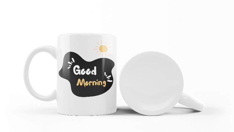 A glossy white ceramic mug with a mountain path design and a text 'GOOD MORNING' on the other side, placed against a plain background.