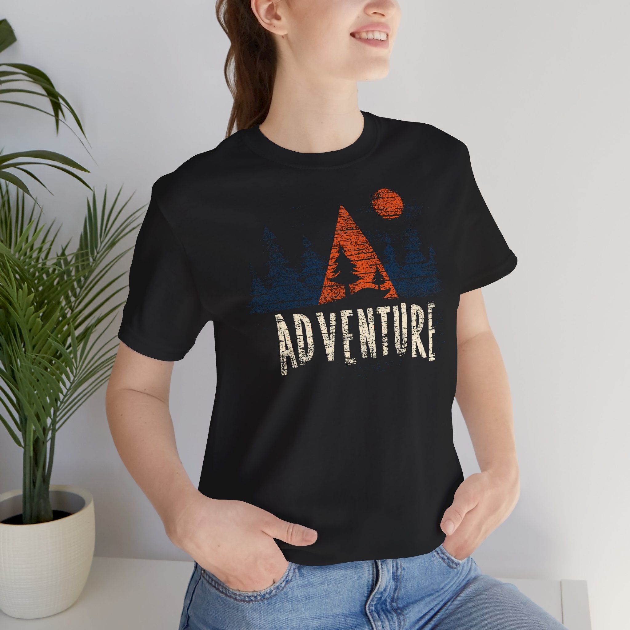 Person wearing a black t-shirt with 'Adventure' and mountain design, standing indoors.
