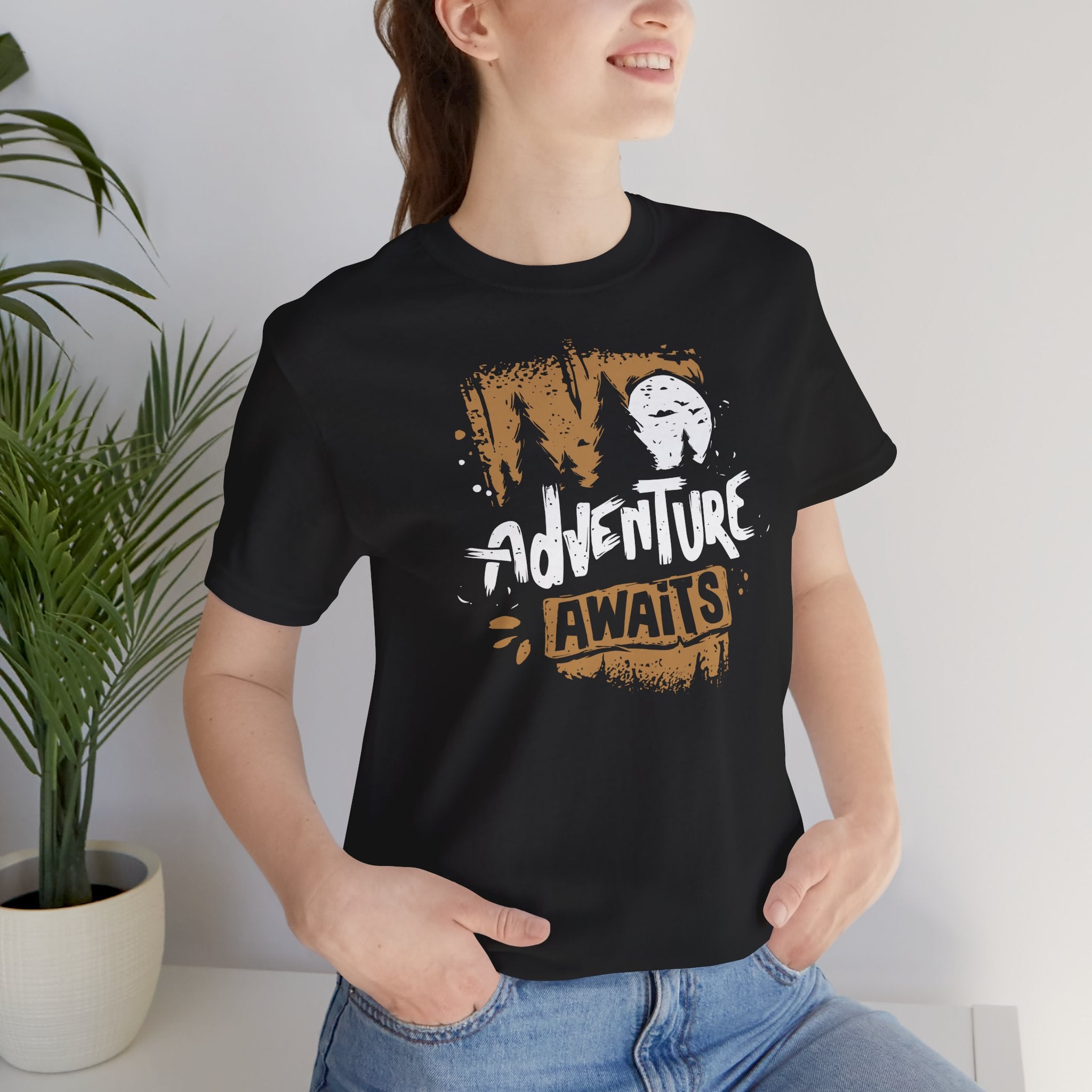 Person wearing a black t-shirt with 'Adventure Awaits' design, standing indoors next to a plant.