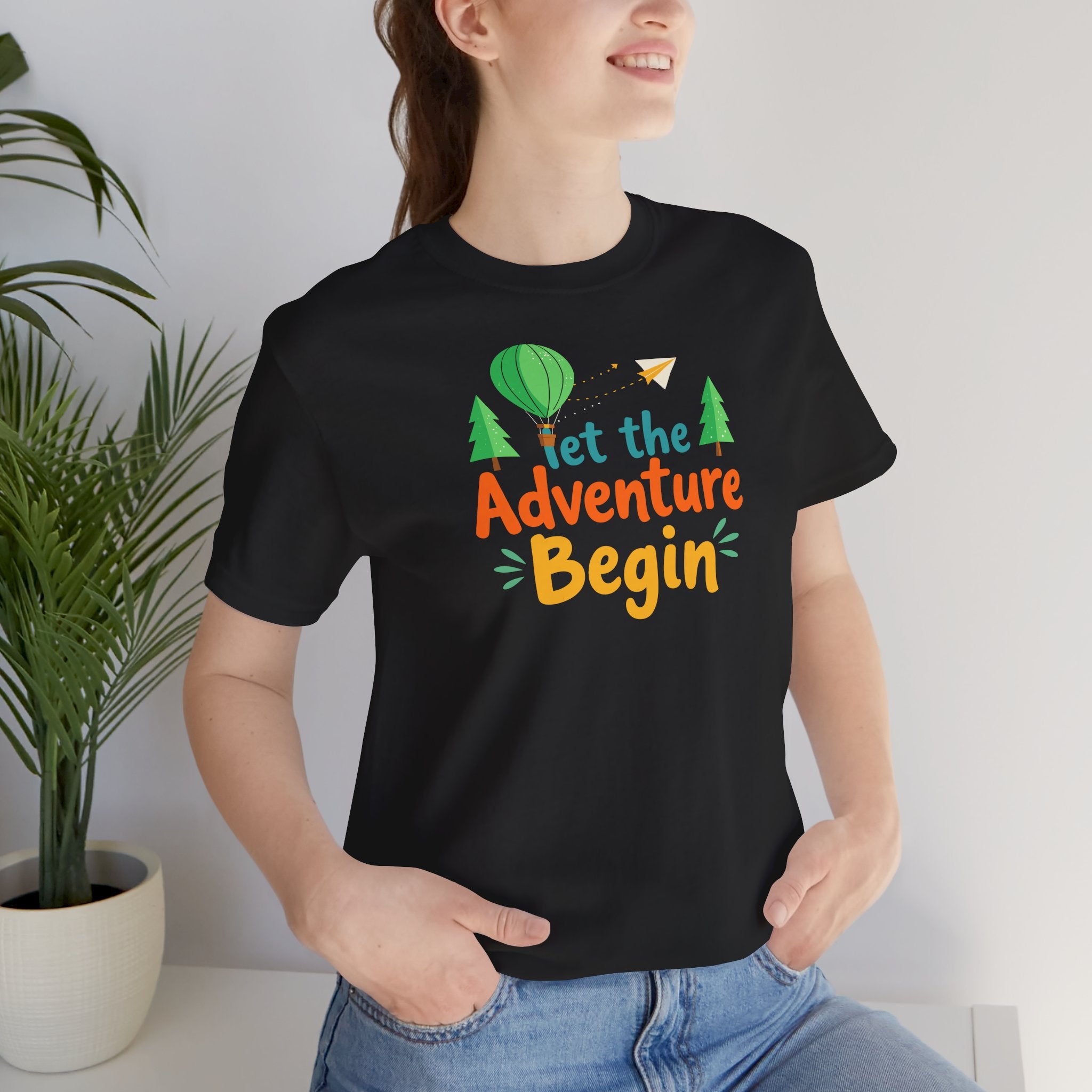 A black crew neck t-shirt with the phrase 'Let the Adventure Begin' printed in a colorful font, accompanied by images of a hot air balloon, trees, and a paper plane.