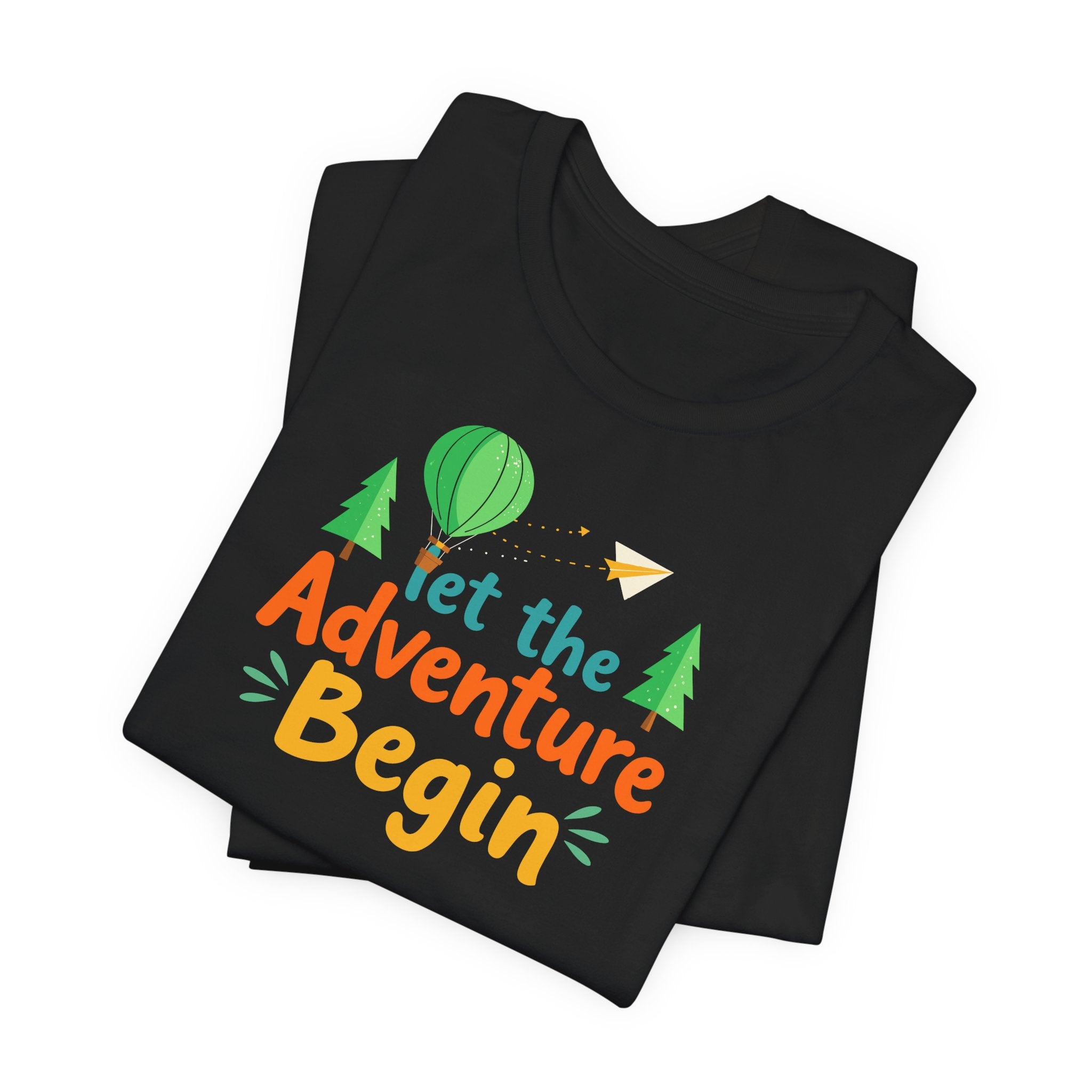 A black crew neck t-shirt with the phrase 'Let the Adventure Begin' printed in a colorful font, accompanied by images of a hot air balloon, trees, and a paper plane.