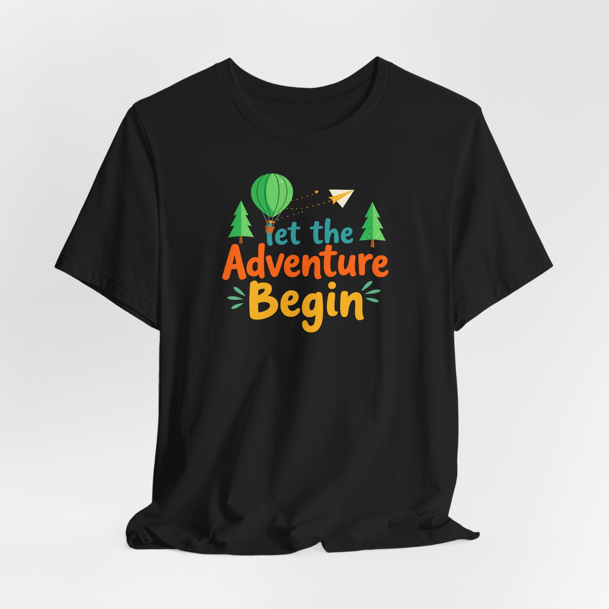A black crew neck t-shirt with the phrase 'Let the Adventure Begin' printed in a colorful font, accompanied by images of a hot air balloon, trees, and a paper plane.