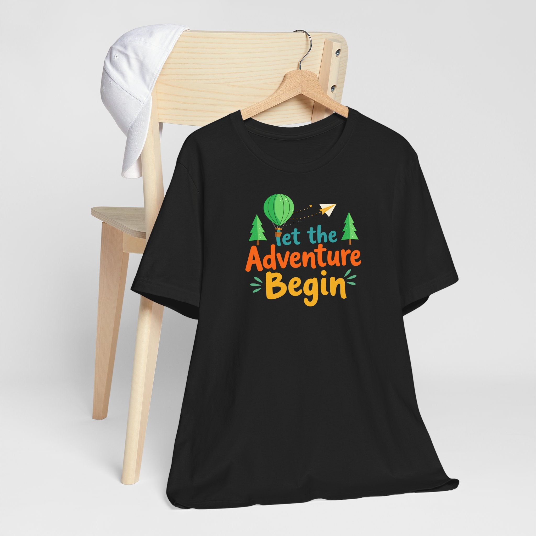 A black crew neck t-shirt with the phrase 'Let the Adventure Begin' printed in a colorful font, accompanied by images of a hot air balloon, trees, and a paper plane.