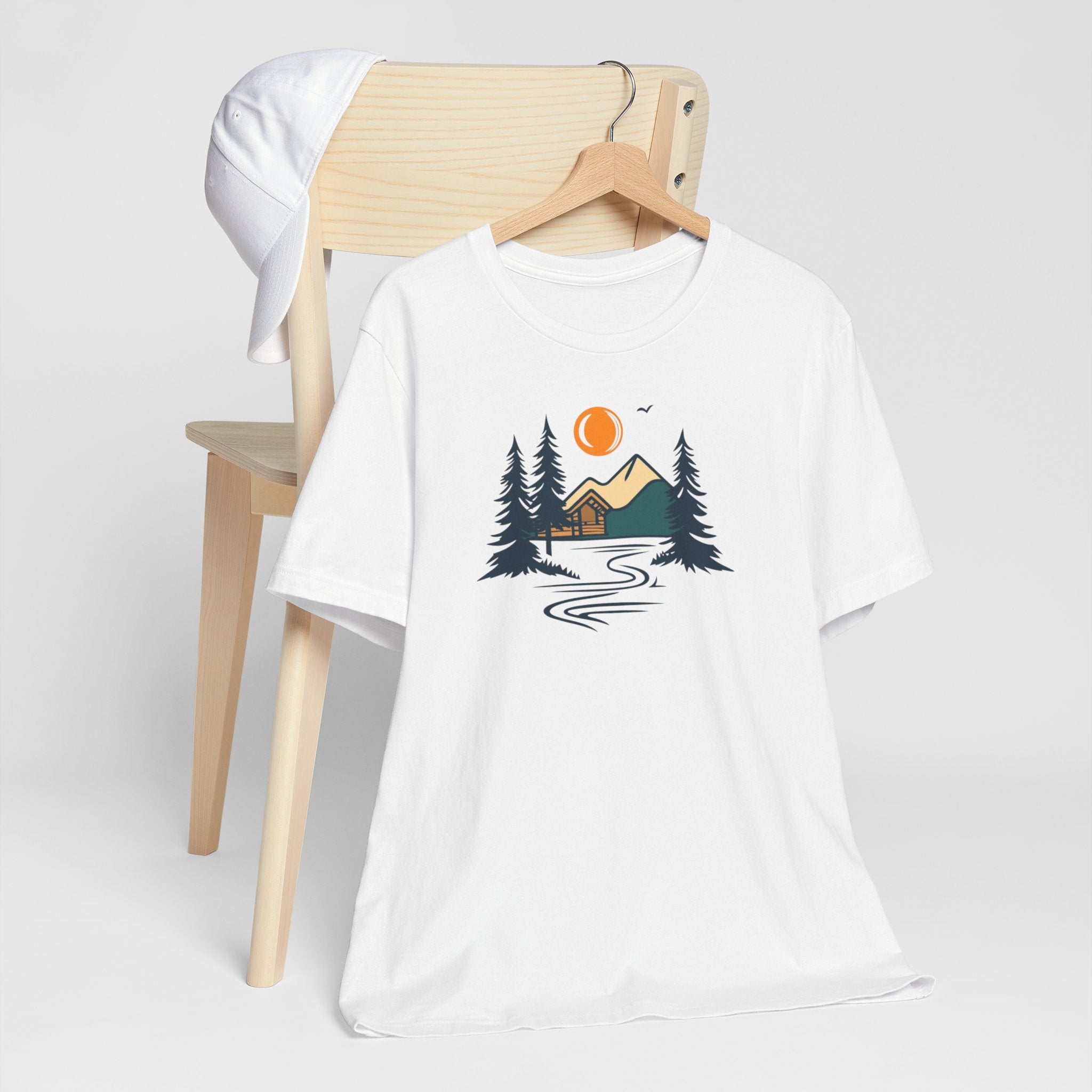 A white cotton t-shirt with a graphic design featuring a cabin, trees, and a mountain range with a warm orange sun.