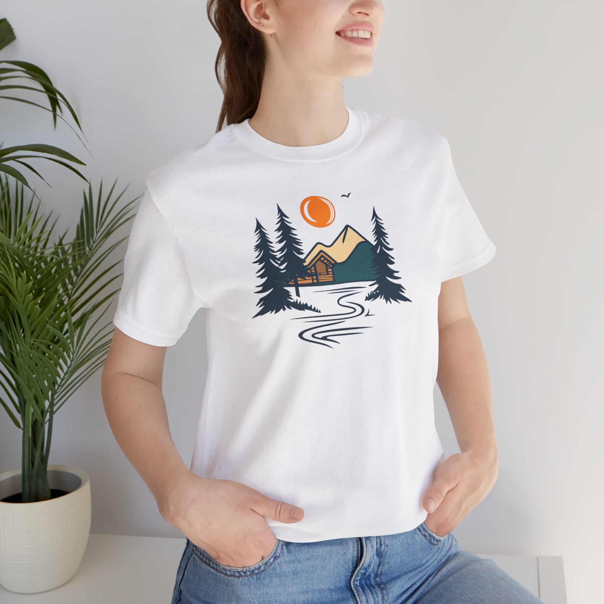 A white cotton t-shirt with a graphic design featuring a cabin, trees, and a mountain range with a warm orange sun.