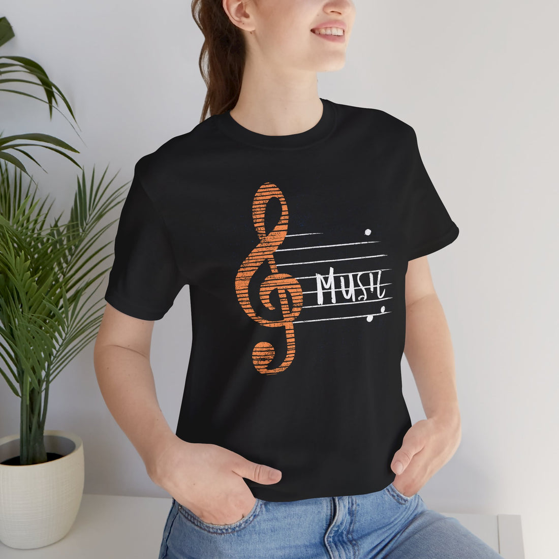Person wearing a black t-shirt with a music-themed design, standing indoors next to a plant.