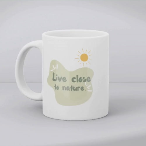 Two white ceramic mugs with a green and brown tree illustration and the quote 'Live Close to Nature'. One mug shows the exterior while the other displays the interior with a beverage.