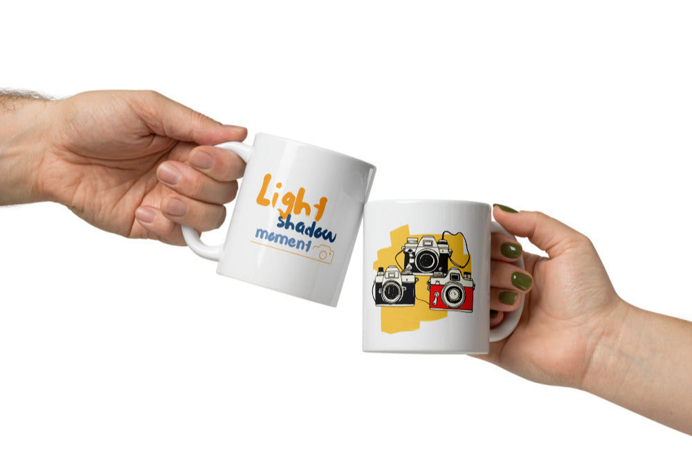 Two ceramic mugs, one with a colorful retro camera collage design and the text 'Light, Shadow, Moment!' The other mug is plain white with no design.